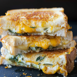 A stack of grilled cheese sandwiches bursting with jalapeños and gooey melted cheese on a dark surface with scattered crumbs.