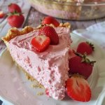 A slice of fluffy strawberry pie on a plate, with fresh strawberries and crumbs in the background.